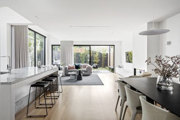 Bright, open-plan contemporary interior combining a white kitchen with a long stone island and black bar stools, a dining area with a dark table, and a living area opening to a landscaped courtyard through extensive glazing. The space features pale timber flooring, white walls and cabinetry, sheer curtains, and a sculptural pendant light.