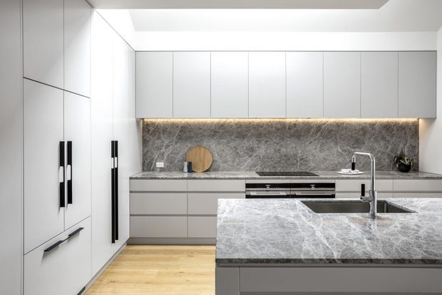 A bright, contemporary galley-style kitchen with extensive pale grey flat-panel cabinetry, tall integrated storage, a large stone island with an undermount sink and streamlined tapware, and a matching stone benchtop and full-height splashback. Warm timber flooring contrasts with the cool grey cabinetry, while concealed under-cabinet lighting adds a refined glow.