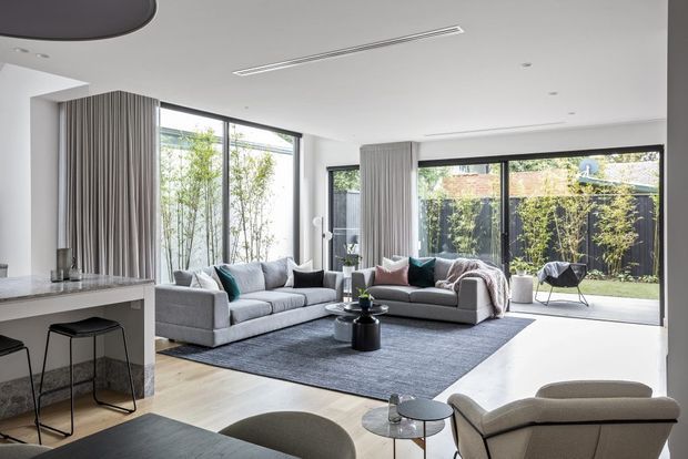 Bright contemporary open-plan living area with a large grey sectional sofa arrangement, dark textured rug, sculptural lounge chairs, floor-to-ceiling glazing, and layered full-height curtains. A stone-topped kitchen island with black stools is visible at left, while the glazing opens toward a planted outdoor courtyard. The space has a clean white ceiling with recessed lighting and linear air-conditioning vents, pale timber flooring, and a neutral grey palette with teal and blush accent cushions.