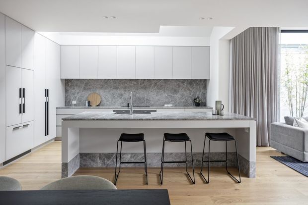A bright contemporary open-plan kitchen with extensive white flat-panel cabinetry, a large central island with seating for three, and a grey veined stone benchtop and full-height splashback. Pale timber flooring, black bar stools and hardware, floor-to-ceiling curtains, and an adjacent living area are visible.