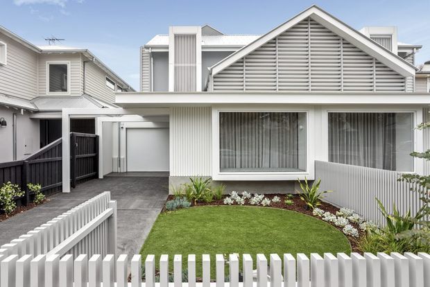 Front exterior view of a contemporary multi-level home with a prominent gabled roof, horizontal louvred gable screening, pale weatherboard-style cladding, white vertical slatted detailing, and large glazed windows. A covered entry and driveway sit beside a compact landscaped front lawn enclosed by a white picket-style fence.