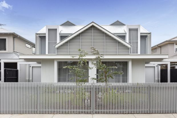 Front exterior view of a contemporary multi-storey residence with a strongly symmetrical composition, white and light-grey vertical cladding, a prominent gabled entry façade with horizontal slatted screening, covered balcony or upper-level form, and a tall pale picket-style privacy fence. Neighbouring houses are visible on both sides.