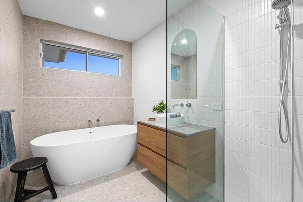 Contemporary bathroom featuring a freestanding white bathtub, walk-in glass shower, wall-mounted timber vanity, large-format pale stone-look wall and floor finishes, and white vertical wall tiles. A narrow horizontal window brings in natural light, while an arched mirrored niche adds visual interest.