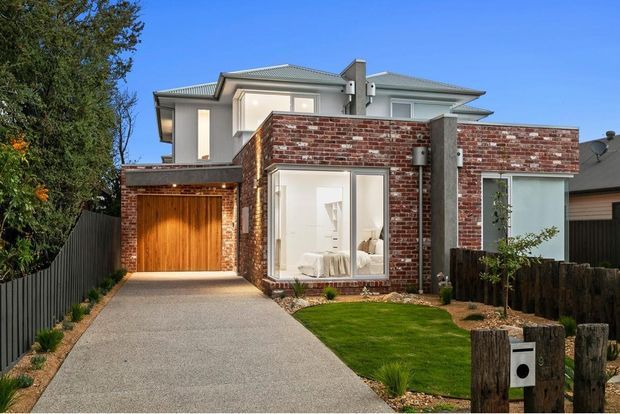 Front exterior view of a contemporary multi-level suburban residence with a prominent red brick facade, pale rendered upper storey, grey metal hipped roof, timber garage door, and a long textured driveway. The compact front garden includes lawn, low planting, timber post fencing, and a letterbox.