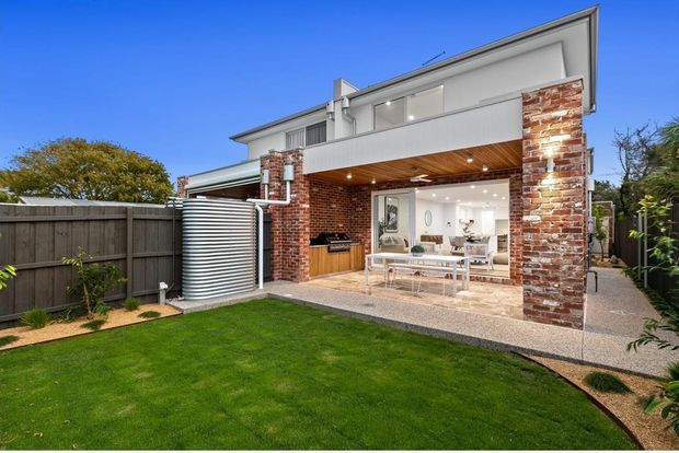 Exterior view of a contemporary two-storey home with a partially covered outdoor entertaining area. The facade combines exposed red brick piers, white rendered upper walls, and a timber-lined soffit. The open ground-level area includes an outdoor kitchen or barbecue zone, dining furniture, and views into a bright interior living/dining space. A large ribbed rainwater tank, fenced lawn, gravel paths, and garden planting are also visible.