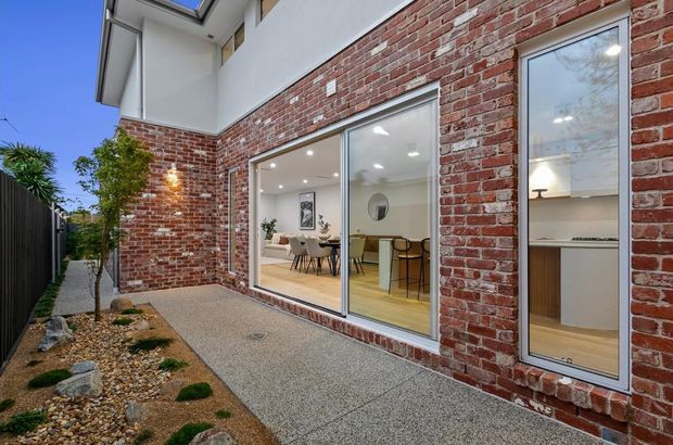 Exterior view of a contemporary brick-clad home, showing a paved side courtyard and large glazed openings. Through the glazing, a bright dining area and a kitchen or built-in joinery are visible. The facade combines exposed red brick with white rendered upper walls and slim aluminium-framed windows and doors.