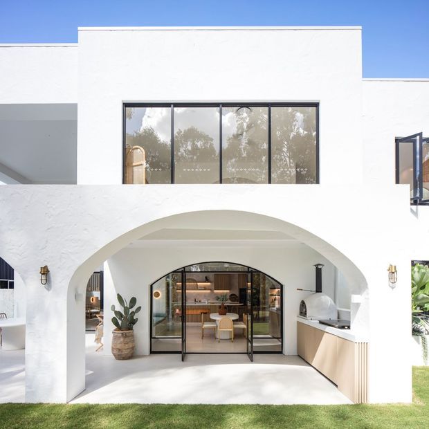Bright white stucco house exterior with a large rounded arch opening over a covered outdoor entertaining area. Black-framed glazing and doors reveal a warm, contemporary interior, while a built-in outdoor kitchen with a barbecue occupies the right side. The facade has a clean, resort-like appearance with strong geometric forms and abundant natural light.