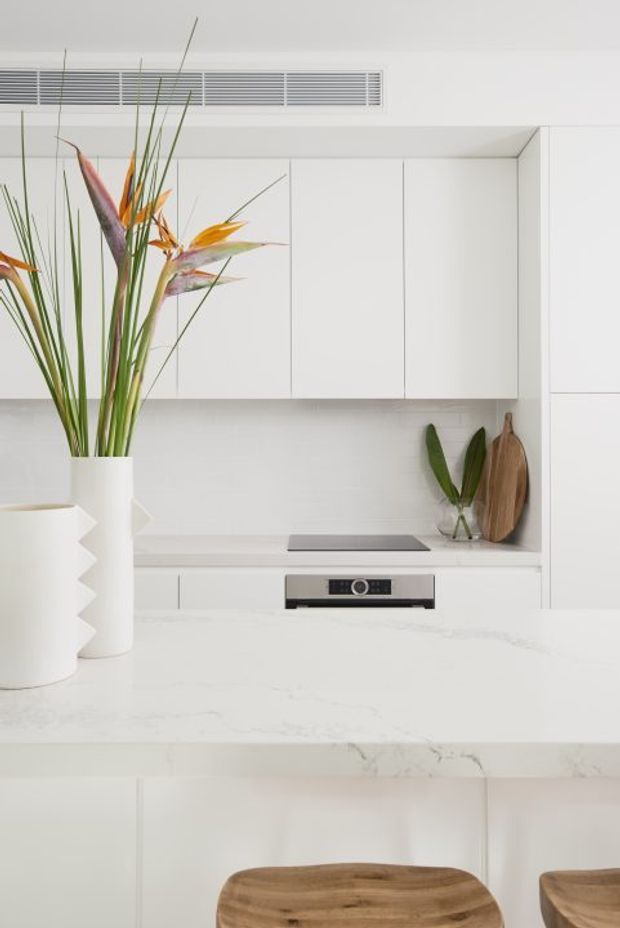 Bright, minimalist white kitchen with flat-panel cabinetry, a white stone island benchtop, integrated appliances and a subtle white tiled backsplash. A sculptural white vase with tropical foliage sits on the island, while timber chopping boards and timber bar stools add warmth.