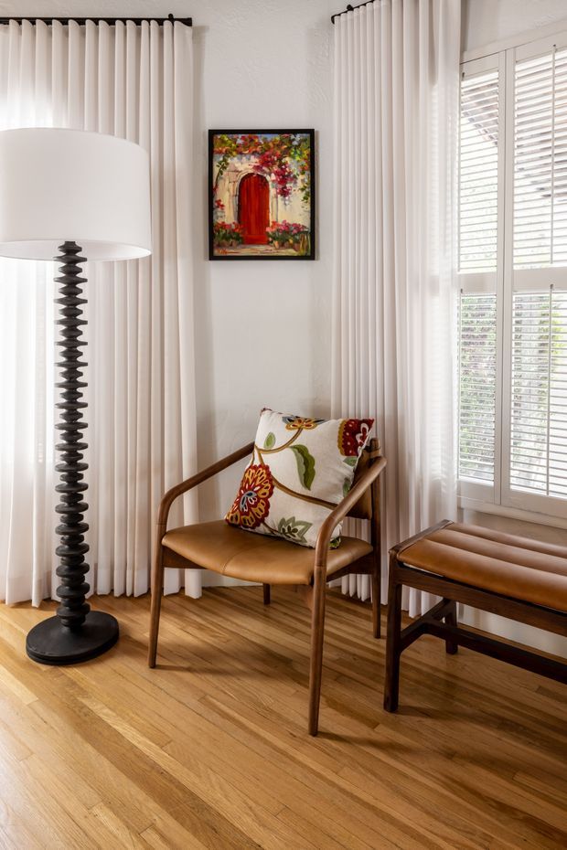 Bright, compact sitting area with warm timber flooring, white floor-to-ceiling curtains, plantation-shuttered windows, and a mid-century style leather armchair with a colourful floral cushion. A matching leather bench and sculptural black floor lamp complete the corner, with colourful artwork on the wall.