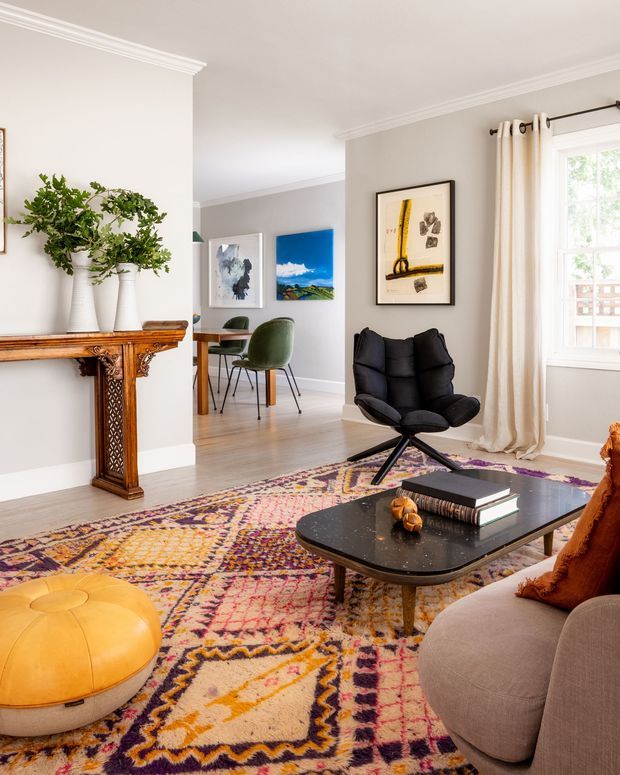 A bright, refined living room with light grey walls, white decorative cornices, timber flooring, and a large colourful patterned rug. The room features a black sculptural lounge chair, rounded coffee table, curved pale sofa, mustard leather ottoman, framed artwork, a classic timber mantelpiece with greenery, and large curtained windows. A dining area is visible through the opening in the background.