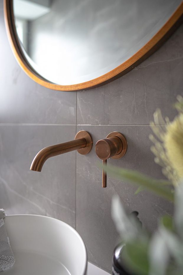 Close-up view of a refined bathroom vanity featuring a wall-mounted brushed bronze/copper-toned basin faucet and matching control on large grey stone-look wall tiles. A white vessel basin, oval timber-framed mirror, and soft greenery are visible, creating a warm contemporary bathroom composition.