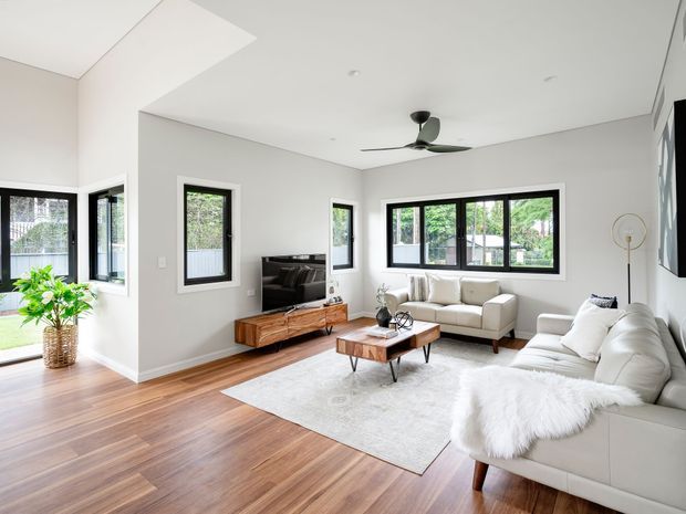 Bright contemporary living room with white walls, extensive black-framed windows, warm timber flooring, and two white sofas arranged around a wood-and-metal coffee table. A timber media console, television, ceiling fan, floor lamp, indoor plant, and large pale rug complete the open, airy space.
