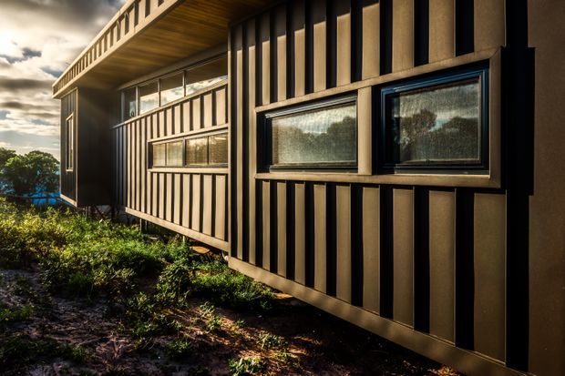 Exterior close-up of a contemporary building with dark vertical ribbed cladding, projecting black-framed horizontal windows, and a covered upper-level balcony or walkway. The structure sits amid planted landscaping and is photographed in dramatic low sunlight.