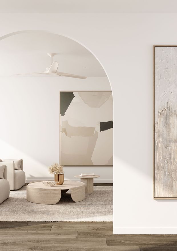 A softly lit contemporary living room viewed through a large rounded archway. The space features warm white walls, a white ceiling fan, neutral upholstered seating, abstract artwork, sculptural pale stone or plaster coffee tables, a textured area rug, and wide-plank timber flooring. The overall composition is calm, minimalist, and tonal, with strong curved architectural detailing.