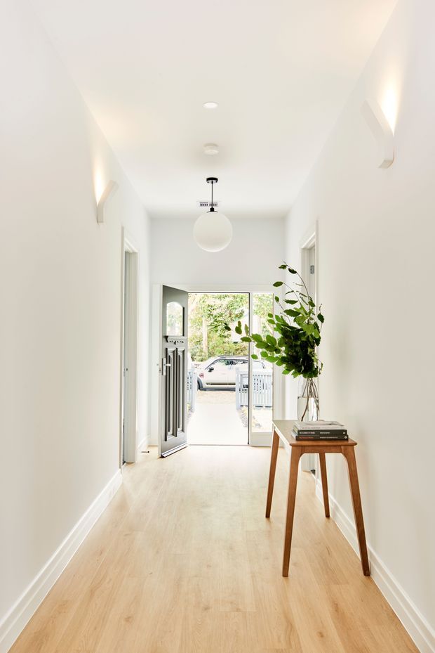 Bright, minimalist residential entry hall with pale timber flooring, white walls and ceiling, recessed lighting, and a spherical pendant light. A narrow wooden console table with books and a tall leafy arrangement lines the right wall. The hall opens through glazed double entry doors to the exterior, with additional doorways along both sides.