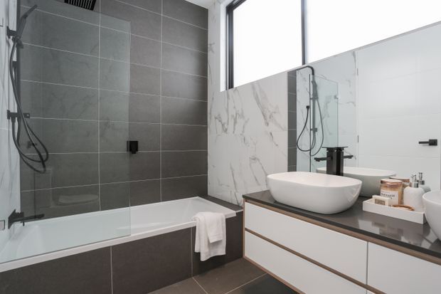 Contemporary bathroom with a tiled bath and shower area behind a frameless glass screen, a freestanding white vessel basin on a dark vanity benchtop, wall-mounted drawers, and a large mirror. The room uses dark grey floor and wall tiles alongside white marble-look wall tiling and black tapware, with natural light from a high window.