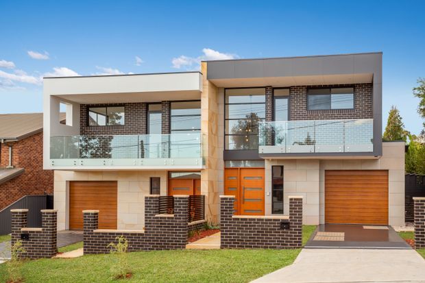 Street-facing view of a contemporary multi-storey residential house with two attached garage-fronted sections, dark brickwork, pale masonry cladding, flat roof forms, extensive glazing, and glass-balustraded upper balconies. Timber garage doors and a bright timber entry door provide warm accents, with low brick boundary walls and a concrete driveway in front.