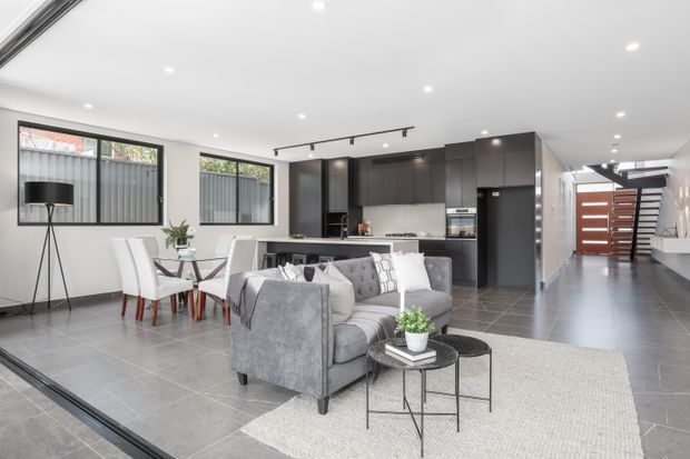 Open-plan contemporary interior combining a living room, dining area, and kitchen. The space features a large grey sectional sofa, white upholstered dining chairs, dark charcoal kitchen cabinetry, grey tiled flooring, recessed ceiling lights, and black track lighting. Full-height glazing and a hallway leading to a modern timber-and-glass entry door are visible.