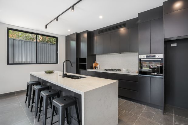 Modern kitchen with a large pale stone island and waterfall end, four black bar stools, matte dark cabinetry, integrated appliances, a grey stone-look splashback, and dark grey tiled flooring. A black-framed window and track lighting contribute to the clean, contemporary fit-out.