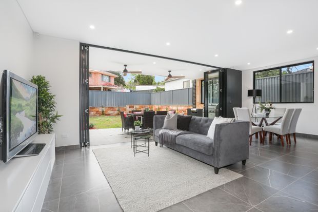 Bright open-plan living and dining room with a contemporary monochrome fit-out. The room features large grey floor tiles, white walls and ceiling with recessed downlights, a grey sectional sofa, white media console, and a dining table with upholstered chairs. Wide black-framed bi-fold or stacking doors open directly to a paved outdoor entertaining area and garden, creating strong indoor-outdoor connection. A dark-framed horizontal window and black feature elements add contrast.