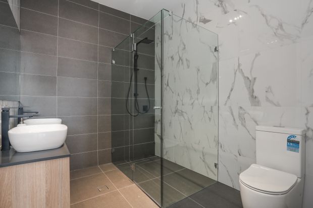 Contemporary bathroom with a floating timber-look vanity and white vessel basin at left, a frameless glass walk-in shower with dark fixtures in the centre, and a wall-mounted toilet at right. Large dark grey wall and floor tiles contrast with a dramatic white marble-look feature wall and bright recessed ceiling lighting.