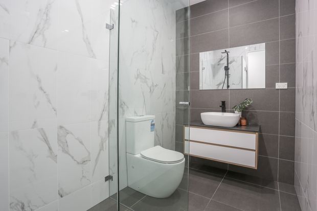 Contemporary bathroom featuring a wall-hung toilet, frameless glass shower enclosure, floating vanity with a white vessel basin, and black tapware. Large white marble-look wall tiles contrast with dark grey floor and feature wall tiles, while a mirror and small plant add practical and decorative detail.