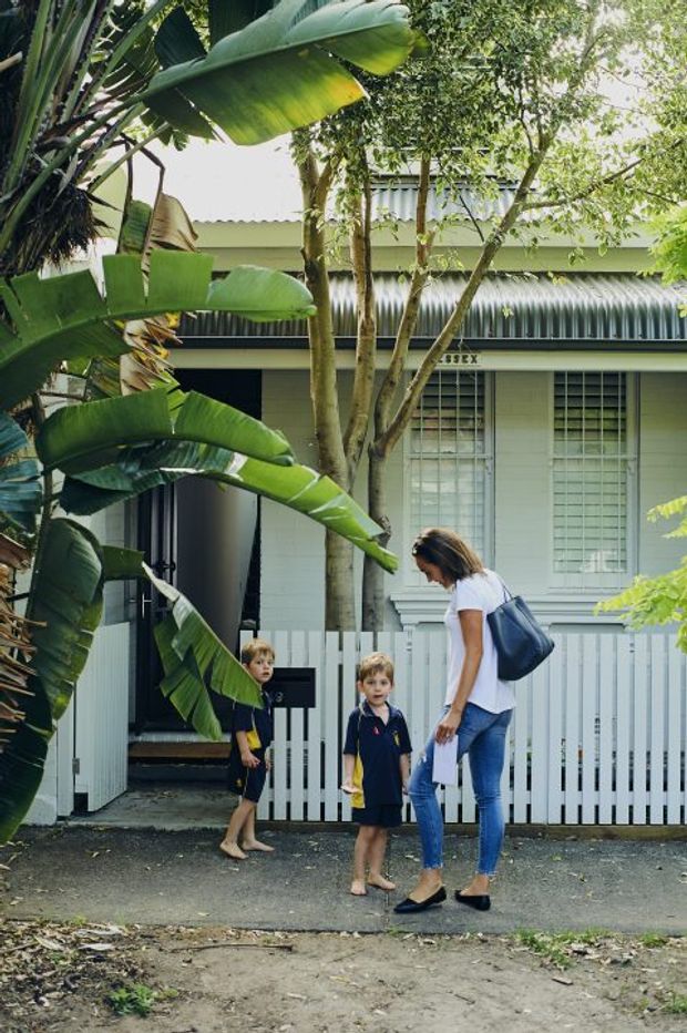Photograph of a leafy residential streetscape showing a compact single-storey house behind a white picket fence. The home has pale painted horizontal cladding, corrugated metal roofing and awnings, security screens, and a small front entry. Tropical foliage, including large banana leaves, frames the scene while an adult and two children stand near the gate.
