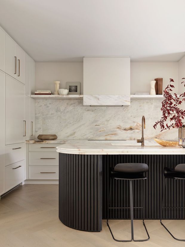A refined contemporary kitchen featuring pale built-in cabinetry, a full-height veined stone splashback, and a sculptural island with a rounded white stone benchtop and vertically fluted dark cabinetry. Two dark bar stools sit at the island, while open shelving displays restrained decorative objects. The space has warm timber flooring, integrated appliances, and soft neutral lighting.