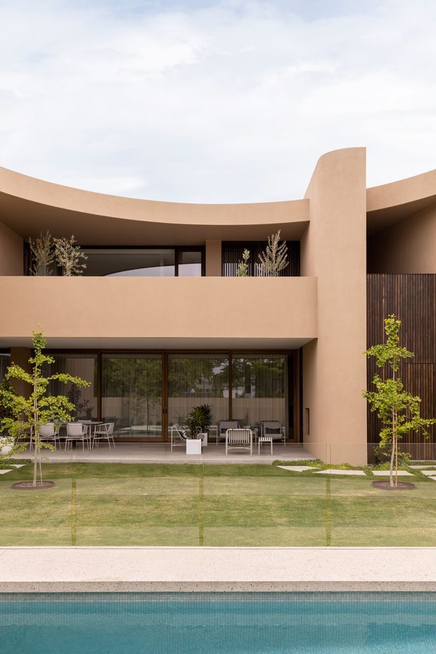 Front-facing exterior view of a contemporary two-storey residence with a curved parapet roofline, warm terracotta stucco walls, dark timber-toned vertical screening, and extensive floor-to-ceiling glazing. A landscaped lawn and patio with outdoor seating sit in front of the house, overlooking a swimming pool in the foreground.