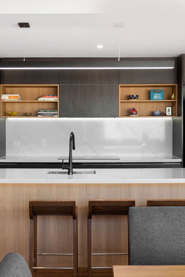 A contemporary kitchen with a large island and integrated breakfast bar, featuring dark wood cabinetry, open timber display shelving, a white veined stone backsplash and benchtop, a black gooseneck tap, and understated linear pendant lighting. Timber bar stools and grey upholstered seating are visible in the foreground.