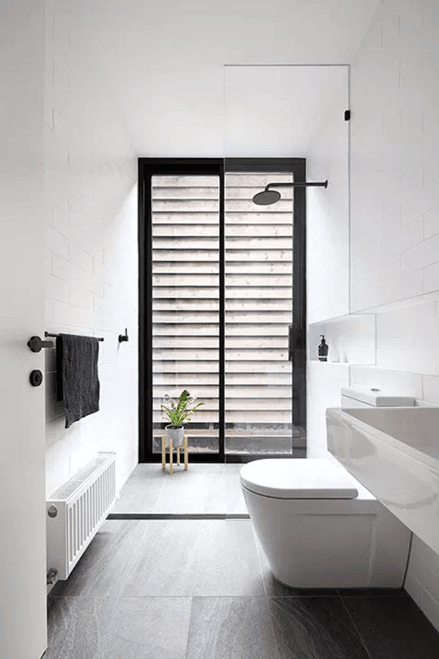 A bright, narrow contemporary bathroom with white tiled walls, pale grey floor tiles, and a black-framed glass shower enclosure at the far end. The shower features a dark wall-mounted rainhead and a textured slatted privacy screen. A wall-hung white toilet sits beside a white built-in vanity with an integrated basin. A black towel rail, dark towel, small plant, and white radiator add contrast. The space is clean, minimalist, and monochromatic.