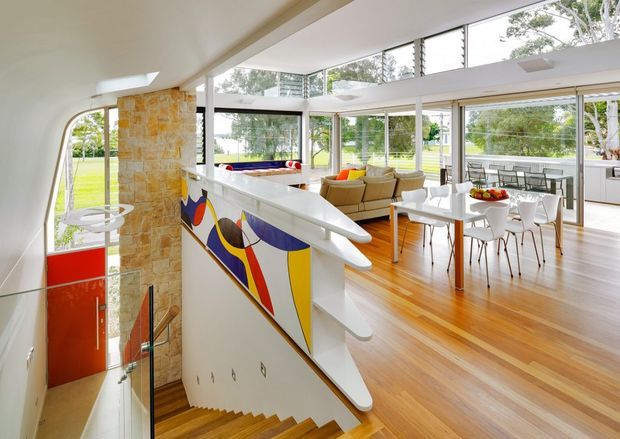 A bright, open-plan mid-century-inspired interior viewed from a stair landing. A timber staircase with glass balustrade descends beside a sculptural white stair railing and a colourful geometric mural panel. The open living and dining area beyond features expansive floor-to-ceiling glazing, warm timber flooring, a white dining table with chairs, and a neutral sofa. A stone-clad column and extensive garden views reinforce the airy connection to the outdoors.
