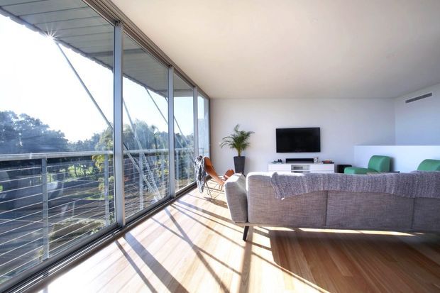 Bright contemporary living room with a large floor-to-ceiling glazed wall opening onto a balcony or terrace. The room features a timber floor, white walls and ceiling, a grey sectional sofa, wall-mounted television, low media console, and strong natural sunlight creating long shadows. The expansive glazing and open outlook dominate the space.