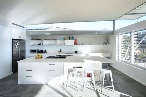 Bright contemporary kitchen with a large white island, integrated white cabinetry, open shelving, stainless-steel appliances, and multiple stools. Full-height glazing and louvred windows bring in abundant daylight, while the dark grey floor contrasts with the predominantly white fit-out.