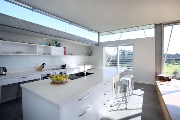 Bright contemporary kitchen with a large white island, integrated cabinetry, open shelving, stainless-steel appliances, and transparent bar stools. Full-height glazing and clerestory windows bring in abundant natural light, with views toward a landscaped outdoor area. A timber-framed raised platform or adjoining space is visible at right.