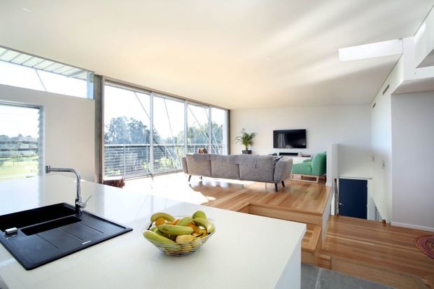 Bright open-plan contemporary interior viewed from a kitchen island, with a white benchtop and integrated black sink in the foreground. The living room features a grey sofa, green armchair, wall-mounted television, timber flooring and expansive floor-to-ceiling glazing opening toward a balcony or exterior view. A split-level arrangement with timber steps is visible on the right.