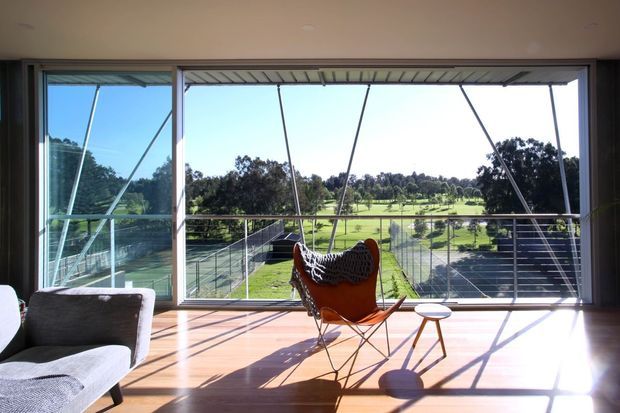 Bright modern living room with floor-to-ceiling glazing opening to a broad green landscape. The room features warm timber flooring, a sculptural leather sling chair with a woven throw, a small timber side table, and a partial upholstered sofa. Strong sunlight and linear window framing create pronounced shadows across the floor.