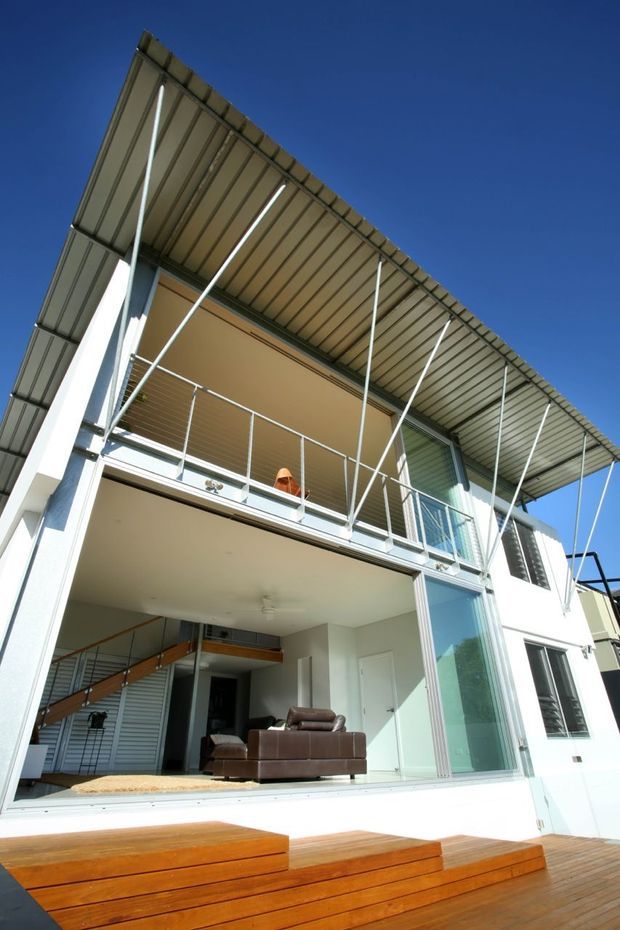 Exterior view of a contemporary multi-storey residence with a prominent deep corrugated metal roof canopy, exposed slender supports, upper balcony, extensive glazing, and a timber deck with broad stepped seating. The open ground level reveals a living area and internal stair.