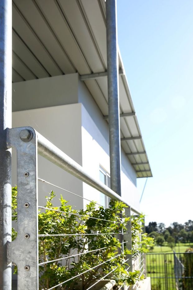 Exterior detail of a contemporary light-coloured building with a shallow corrugated metal roof, exposed metal support posts, and a galvanized steel wire-and-post railing enclosing a planted edge. Bright daylight and a green suburban landscape are visible beyond.