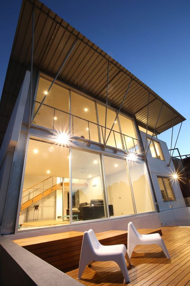 Evening exterior view of a contemporary multi-level house with extensive floor-to-ceiling glazing, a projecting timber-lined roof canopy supported by slender steel braces, and a timber deck with white outdoor chairs. Warm interior lighting reveals an open-plan living area and stairway behind the glass.