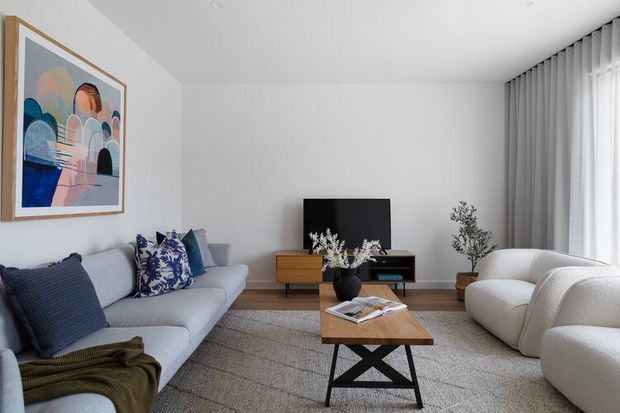 A bright, contemporary living room with a low light-grey sectional sofa, sculptural cream lounge chairs, a timber coffee table with black crossed legs, a simple media console, and a large abstract artwork. Full-height sheer curtains provide soft daylight, while a textured neutral rug, blue patterned cushions, greenery, and a dark green throw add restrained colour and texture.