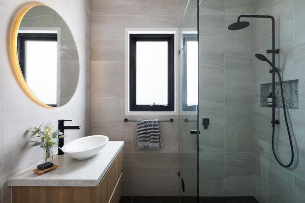 Contemporary bathroom with a floating timber vanity, pale stone benchtop, vessel basin, matte black tapware, large round backlit mirror, and a frameless glass walk-in shower. Large-format light grey wall tiles and dark floor tiles create a refined, neutral scheme, with black-framed windows providing natural light.