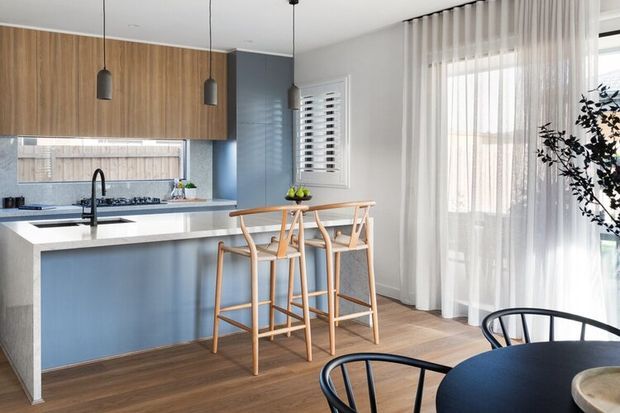 Bright contemporary open-plan kitchen and dining area with a blue-grey island, white stone benchtop, warm timber cabinetry and flooring, pendant lighting, timber bar stools, and sheer floor-to-ceiling curtains. A compact dining table with black chairs occupies the foreground.