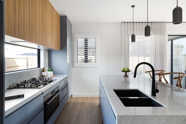 Contemporary kitchen with a long central island, integrated black sink and tap, gas cooktop, built-in oven, dark lower cabinetry, and warm timber overhead cabinetry. The room features a pale stone benchtop, grey-blue cabinetry, timber flooring, sheer curtains, pendant lighting, and abundant natural light.