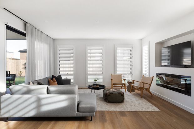 Bright contemporary living room with a large light-grey sectional sofa, timber flooring, plantation shutters, sheer curtains, two timber lounge chairs, a round coffee table, and a built-in media wall with television and fireplace. The room opens toward an outdoor area through full-height glazing.