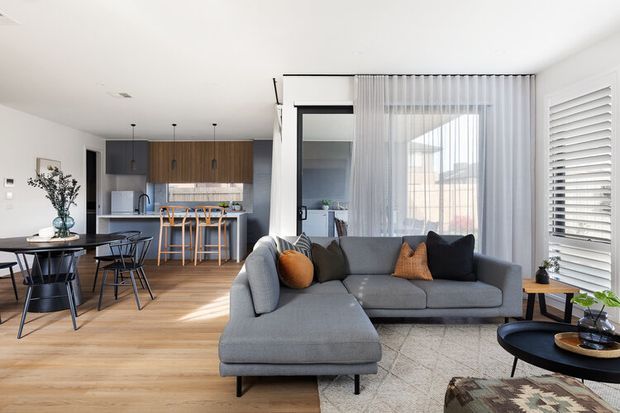 Bright open-plan living and dining area with a contemporary kitchen at the rear. The room features pale timber flooring, white walls, dark kitchen cabinetry with timber overhead joinery, a grey sectional sofa, sheer curtains, a black-framed glazed door, and white plantation shutters. Natural daylight creates an airy, relaxed atmosphere.
