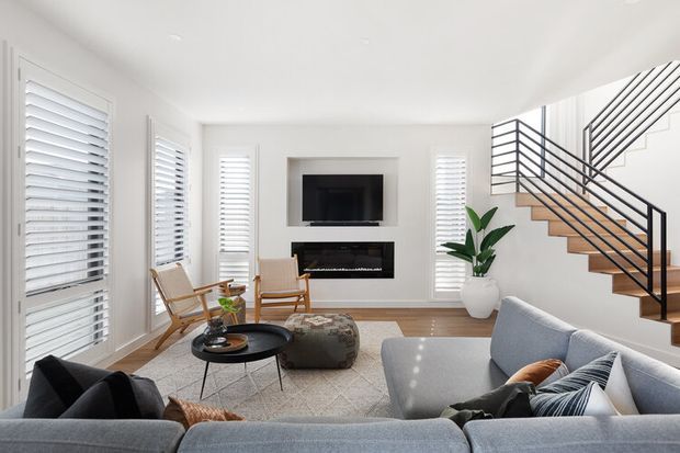 Bright contemporary living room with a large grey sectional sofa, patterned area rug, round black coffee table, timber accent chairs, wall-mounted television and linear fireplace. White plantation shutters line the windows, while an open timber staircase with black metal balustrading rises along the right side. A potted plant adds greenery to the otherwise neutral interior.