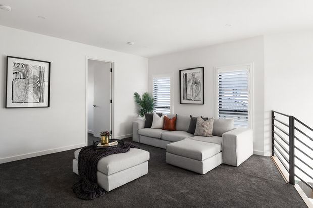 A bright, contemporary living room with a light grey sectional sofa and ottoman, dark charcoal carpet, white walls, plantation shutters, monochrome artwork, and a black metal stair balustrade overlooking the space. A small plant and neutral cushions add warmth and texture.