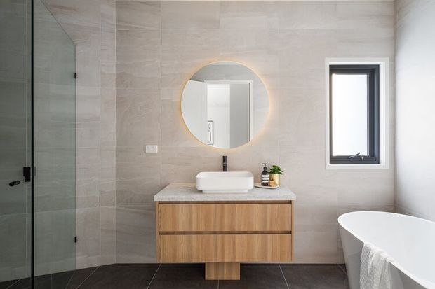 Contemporary bathroom featuring large pale stone-look wall tiles, dark floor tiles, a wall-mounted timber vanity with a white vessel basin, round backlit mirror, frameless glass shower, black tapware, and a freestanding white bathtub. The space is bright and refined with warm timber detailing.