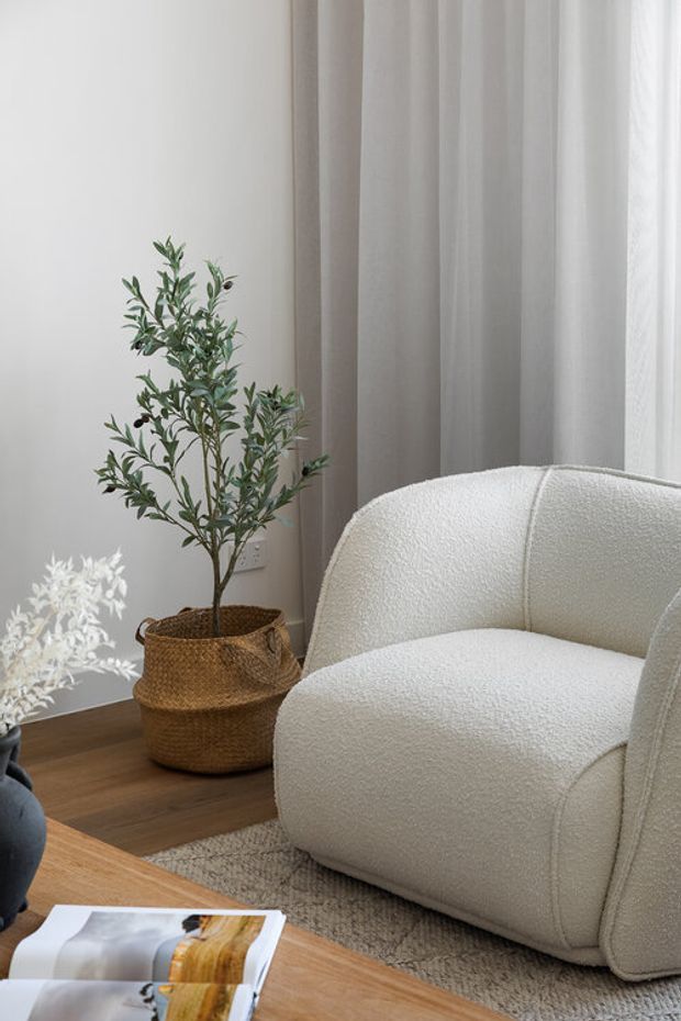 A softly lit, neutral-toned living room corner featuring a large off-white boucle armchair, sheer floor-to-ceiling curtains, warm timber flooring, a textured neutral rug, and a woven basket planter with an indoor tree. A timber coffee table with an open magazine occupies the foreground. The styling is calm, organic, and minimalist.
